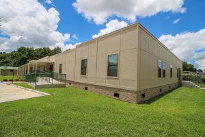 A large-scale modular office complex photographed from the front, featuring beige-toned painted exterior siding. The building has multiple staggered roof lines with a professional architectural design. Dark-tinted windows line the facade. The foreground shows manicured landscaping with evergreen shrubs. Clear blue sky visible above. This demonstrates Rose's capability for multi-unit commercial construction.